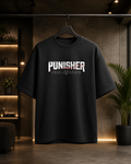 A black short-sleeve graphic T-shirt hanging on a black hanger against a dark, modern interior backdrop with warm shelving and a potted plant. The shirt features the word 'PUNISHER' in large distressed white letters with red and gray distressed elements, and 'ANGEL DEATH' in smaller distressed white letters underneath.